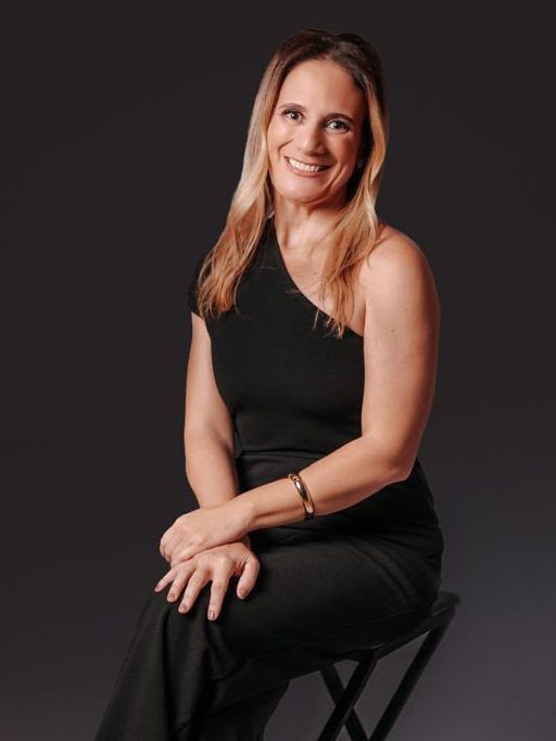 Adriana Trocilo Picanço Rostagno, advogada previdenciarista, sentada em um banco alto, de macacão preto, em fundo de estúdio.