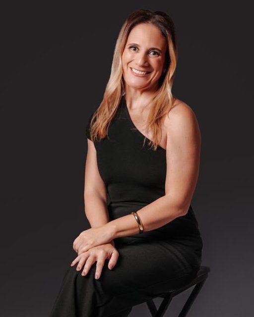 Adriana Trocilo Picanço Rostagno, advogada previdenciarista, sentada em um banco alto, de macacão preto, em fundo de estúdio.